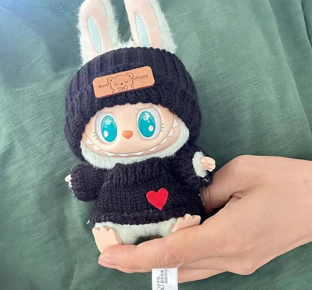 Labubu with a black knit hat and sweater held by a hand on a green fabric background.