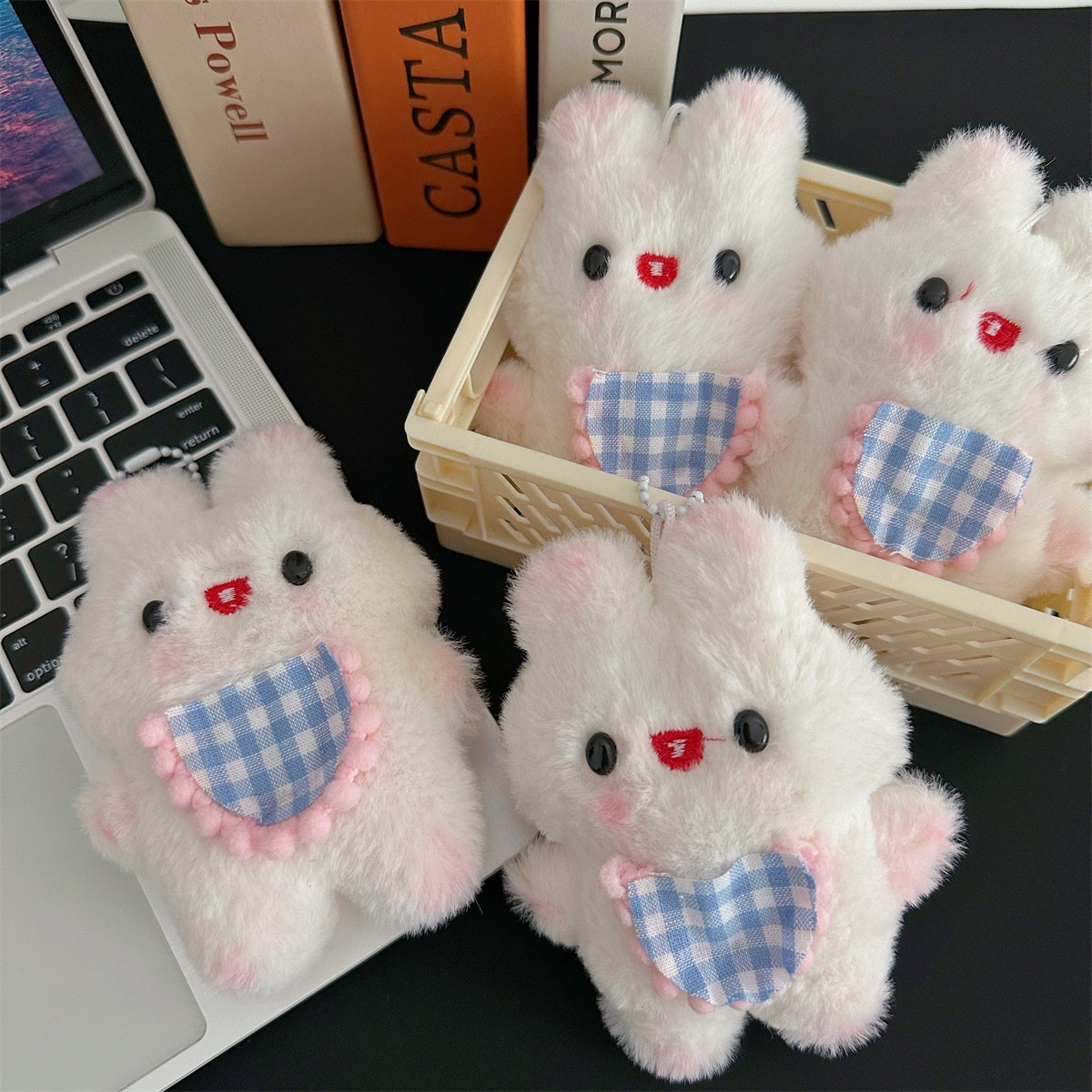 White stuffed animal keychain with blue checkered bibs on a desk next to a laptop