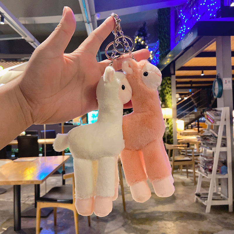 Two plush llama keychains, one white and one pink, held by a hand in an indoor setting.