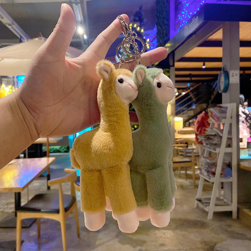 pink and green plush llama keychains held by a hand in an indoor setting with tables and chairs.