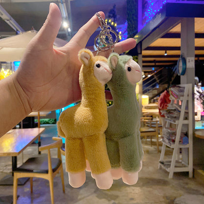 pink and green plush llama keychains held by a hand in an indoor setting with tables and chairs.