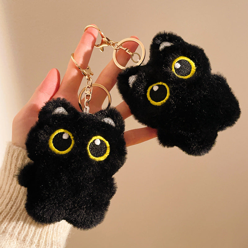 Black fluffy cat-shaped keychains with yellow eyes held by a hand against a beige background