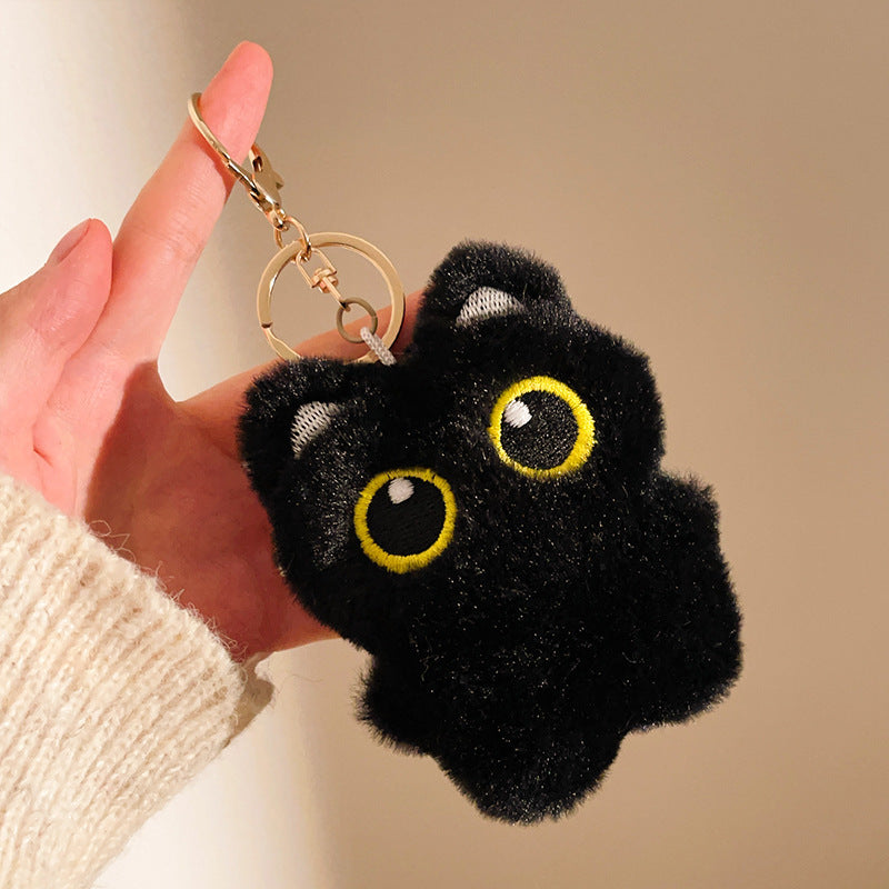 Black plush keychain with yellow eyes held by a hand against a beige background