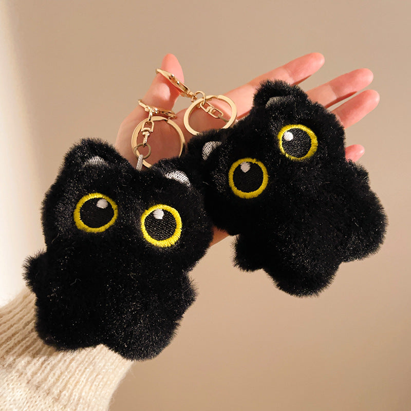 Black plush cat-shaped keychains with yellow eyes held by a hand against a beige background