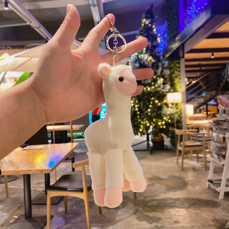 White plush keychain shaped like a llama held by a hand in an indoor setting with tables and chairs.